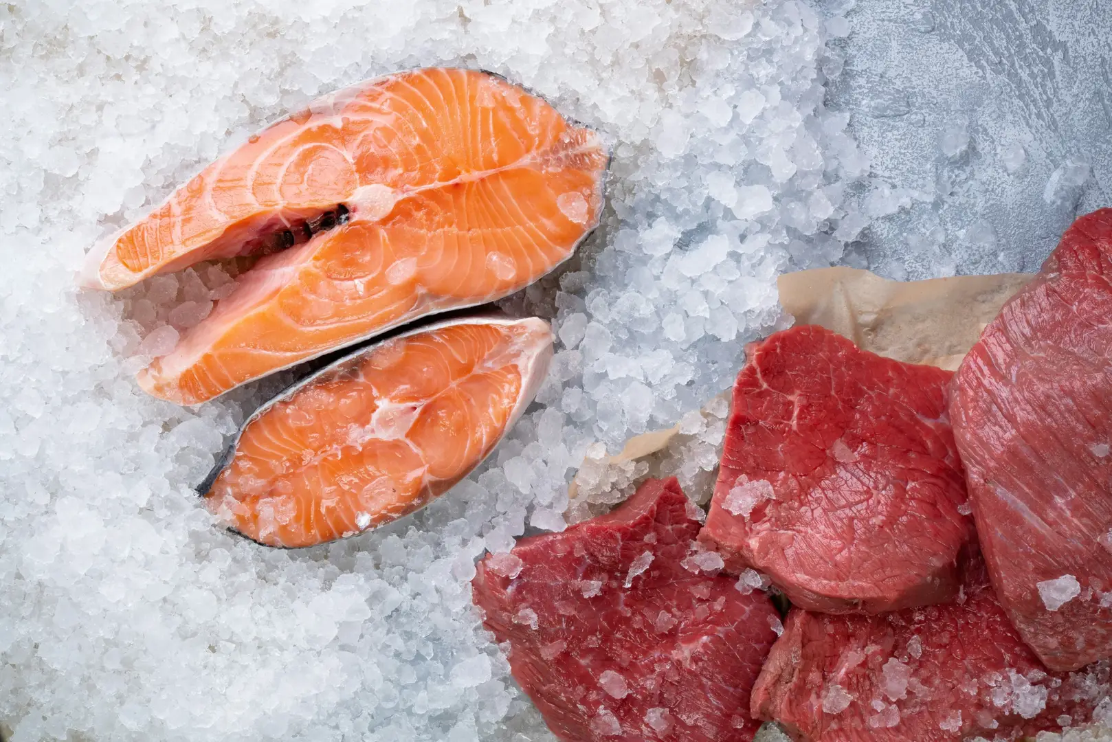 Auf dem Foto sieht man zwei Stücke frischen Fisch, sowie vier Stücke frisches rotes Fleisch, die auf Eis liegen und von diesem Umgeben sind.